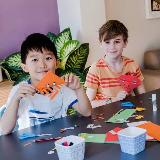 Two boys sat at a table crafting and showing paper bookmarks they've created
