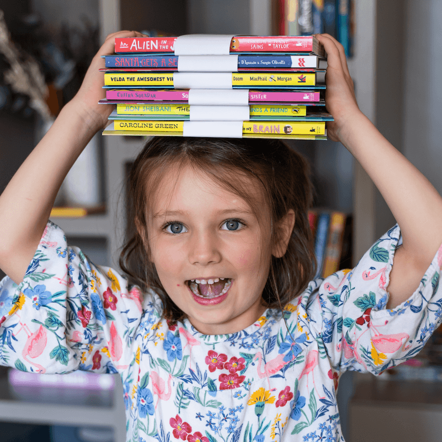A smiling child holding a pile of Parrot Street Book Club book and activity pack bundles on her head.