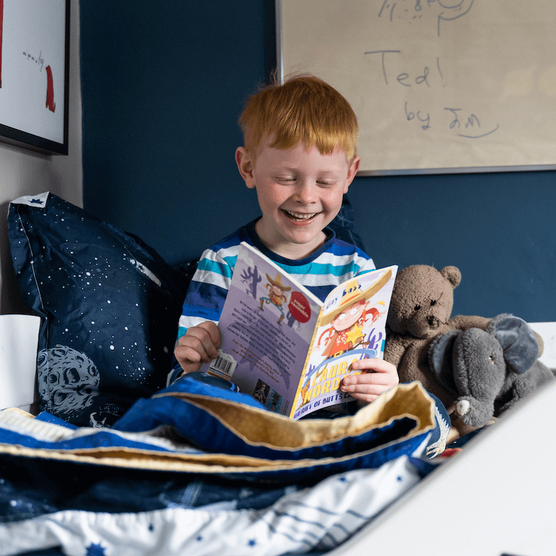 A smiling boy sitting on a bed cross-legged reading a chapter book from a Parrot Street Book Club subscription box