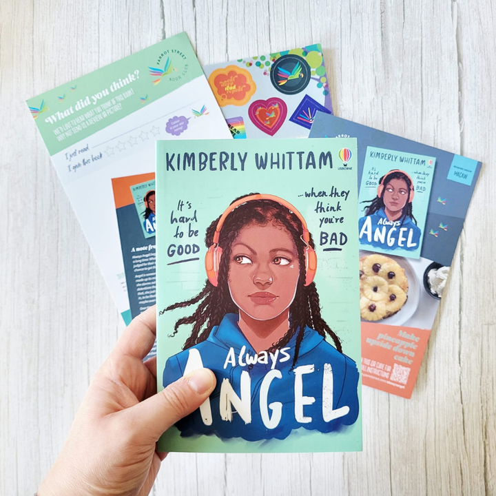 Hand holding a book titled 'Always Angel' by Kimberly Whittam with other books in the background on a wooden surface.