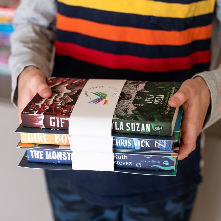 A child's hands holding a pile of three wrapped chapter book and activity bundles from Parrot Street Book Club