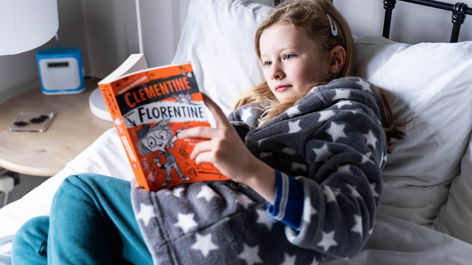 A girl in a dressing gown lies on a bed reading a chapter book, from a Parrot Street Book Club subscription box