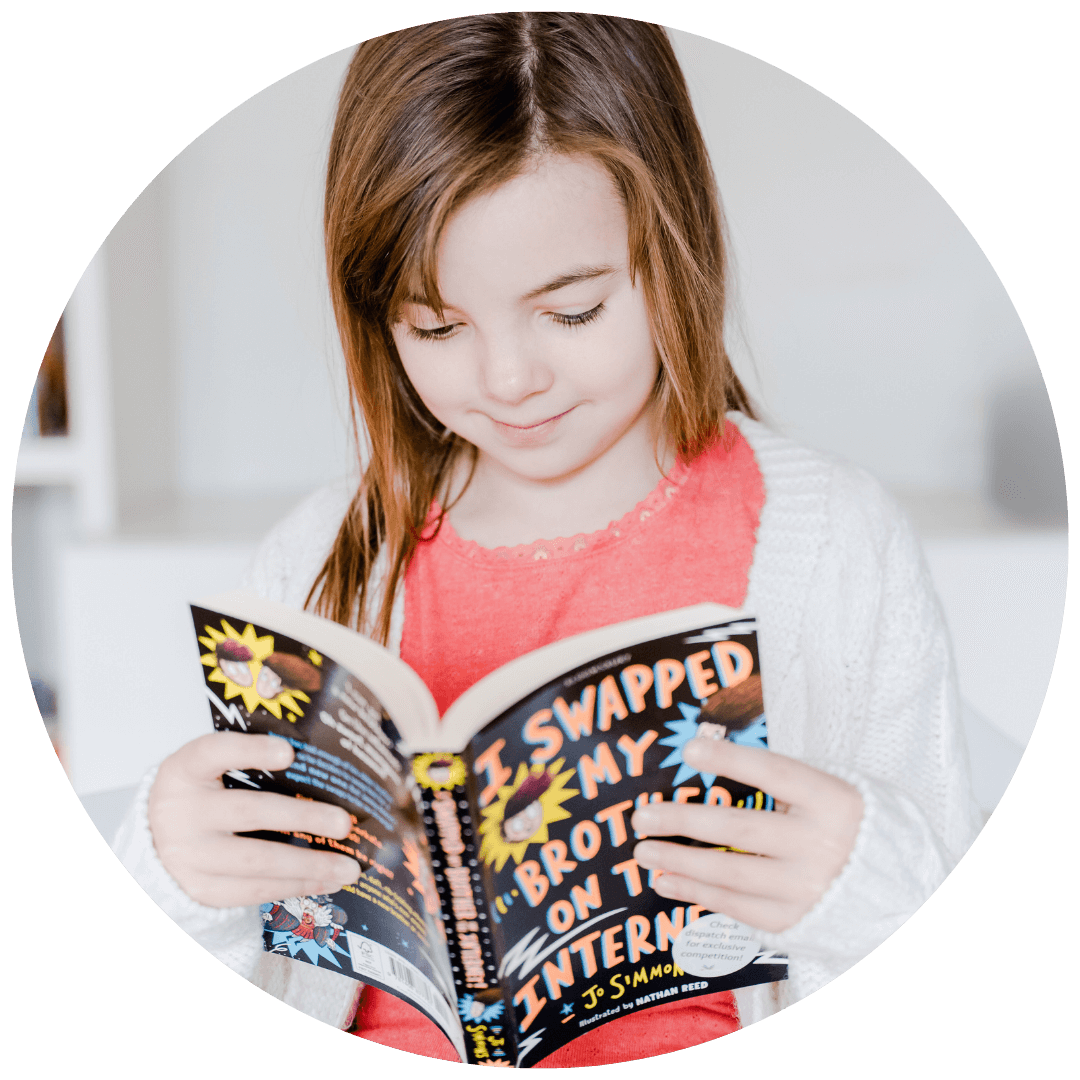 A girl in a pink top looking down and reading a chapter book with black and orange cover
