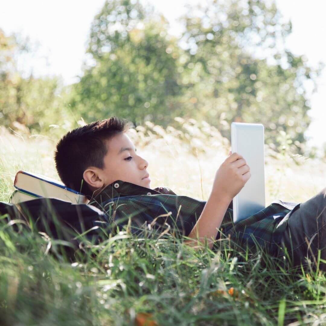 A boy lying in grass reading with head resting on a pile of books