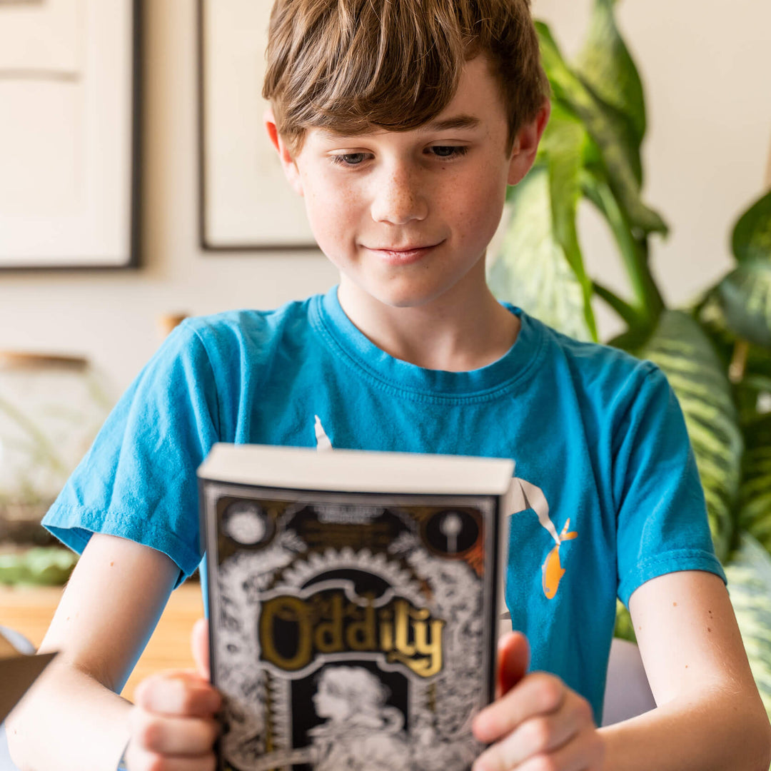 A tween boy holding a substantial chapter book from a Parrot Street Book Club Macaw subscription box