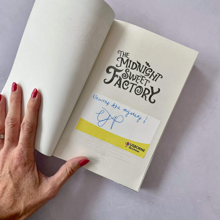 Open book with 'The Midnight Sweet Factory' title, signed bookplate and a hand holding it.