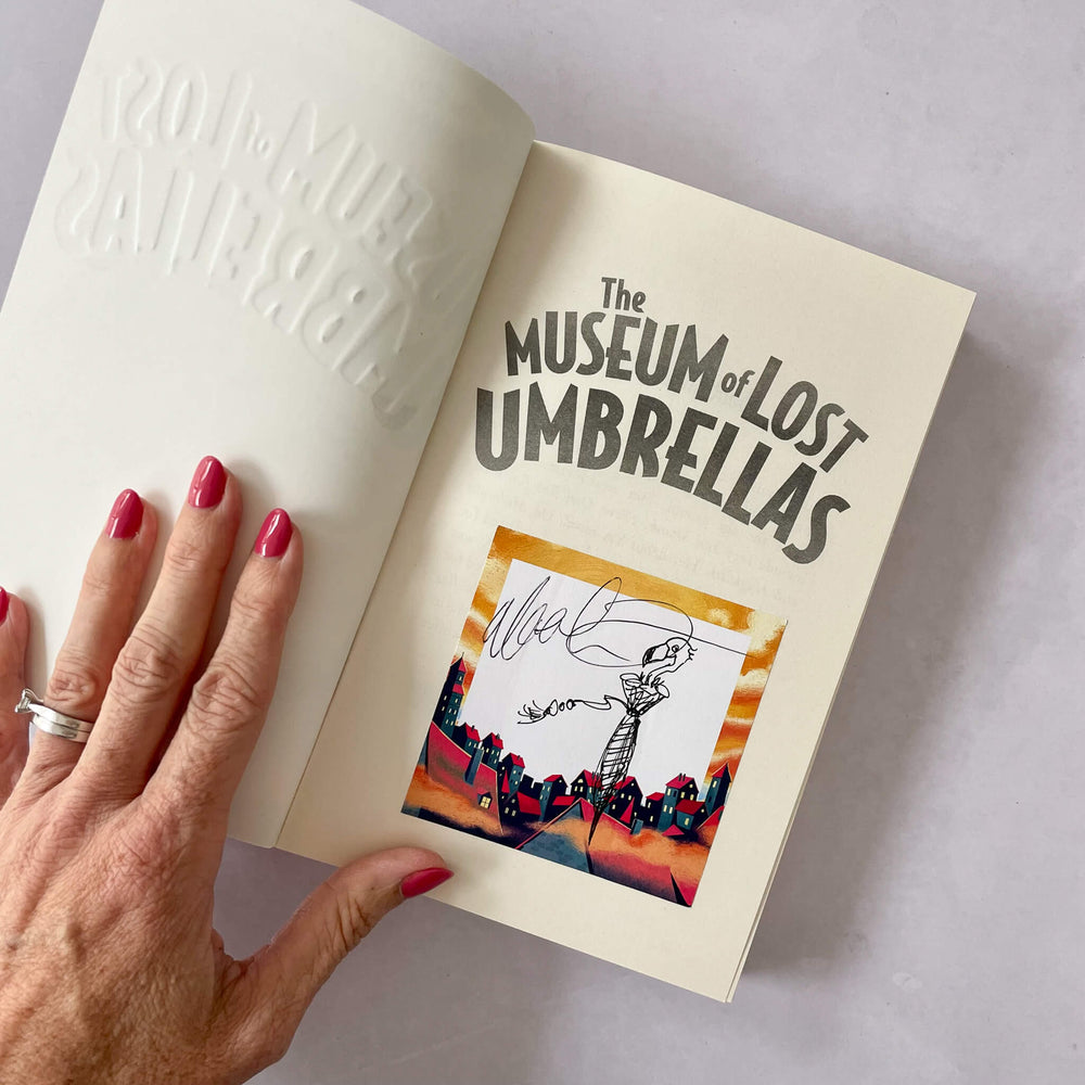 Open book titled 'The Museum of Lost Umbrellas' with signed bookplate and a hand holding it.