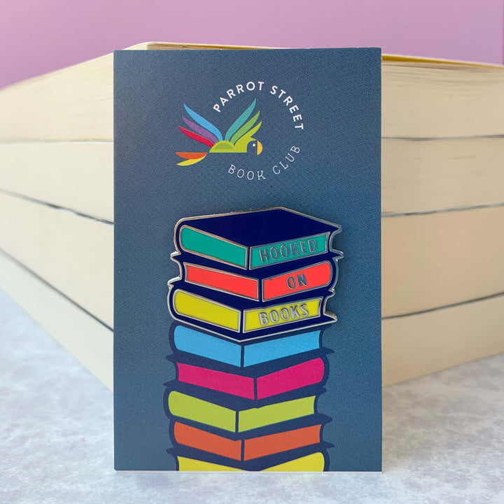 Enamel Hooked on Books pin badge shown on colourful backing card resembling a pile of multicoloured books.