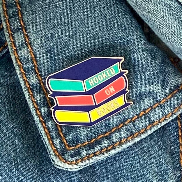 Enamel pin badge shaped like a pile of books reading Hooked on Books. 