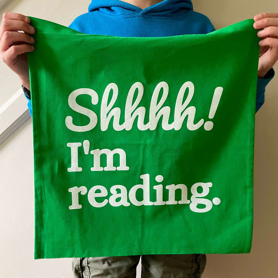 A child holding up a green cotton tote bag with the slogan Shhh! I'm reading. 